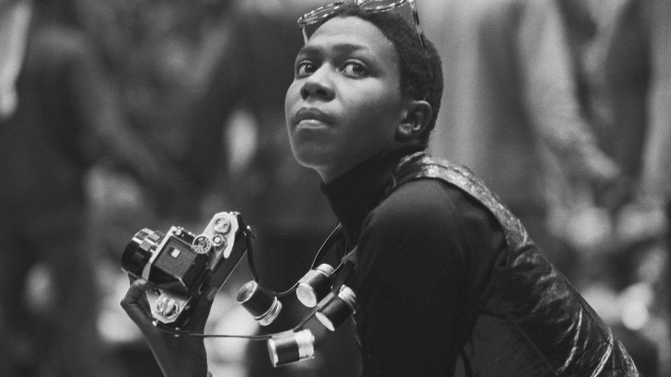 afeni shakur looks to her left off camera in this black and white photo, she is holding a film camera and wears glasses on her head and a turtle neck and vest