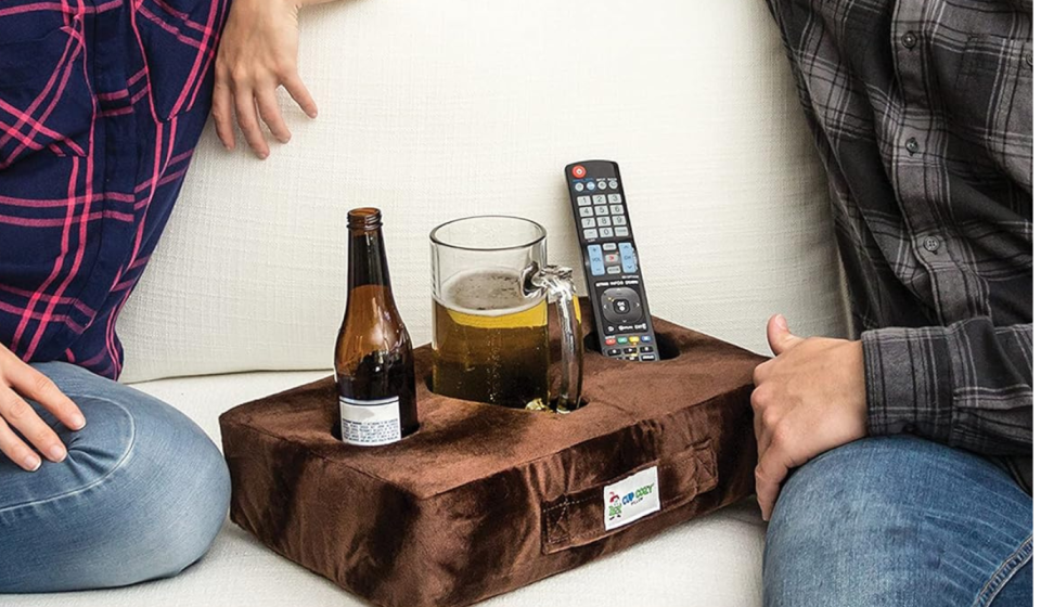 two people on a couch next to the cup pillow, which is holding beer and a remote