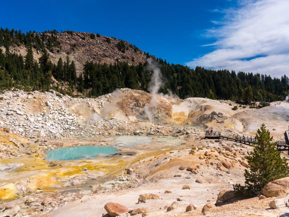 A turquoise pool of water in a hydrothermal area surrounded by orange dirt and rocks. There are also tall evergreen trees.