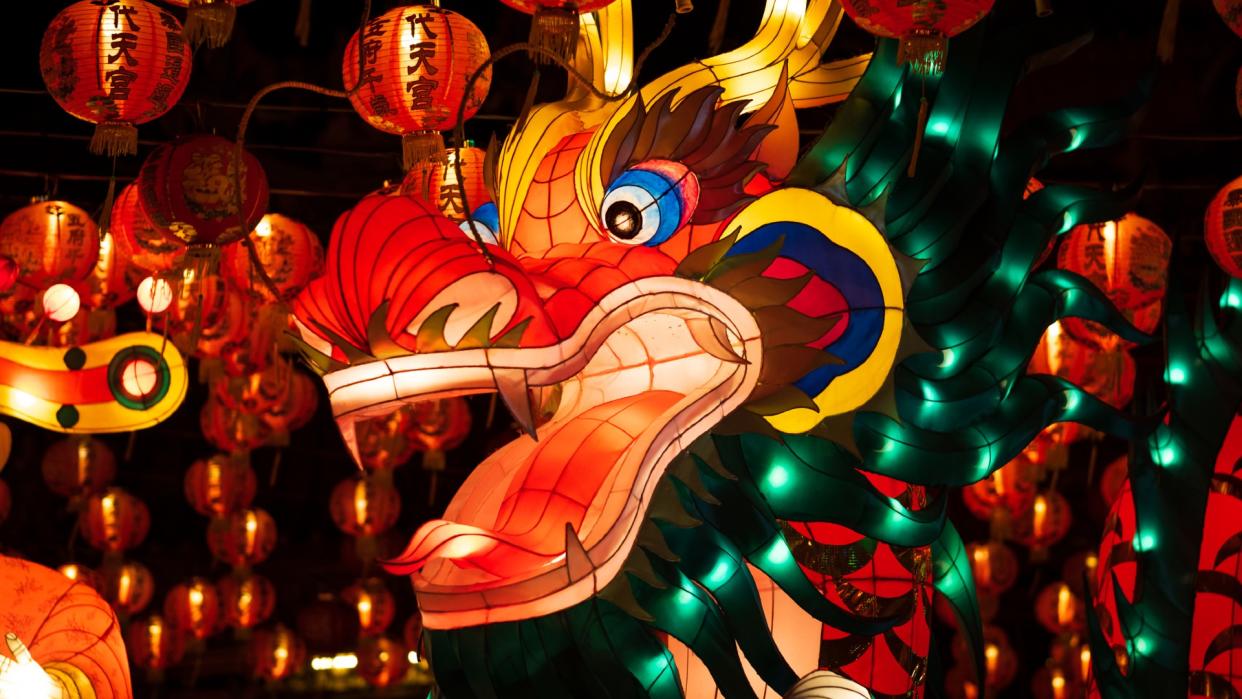  A brightly colored dragon-shaped lantern lit up at night surrounded by red spherical lanterns covered in Chinese characters. 