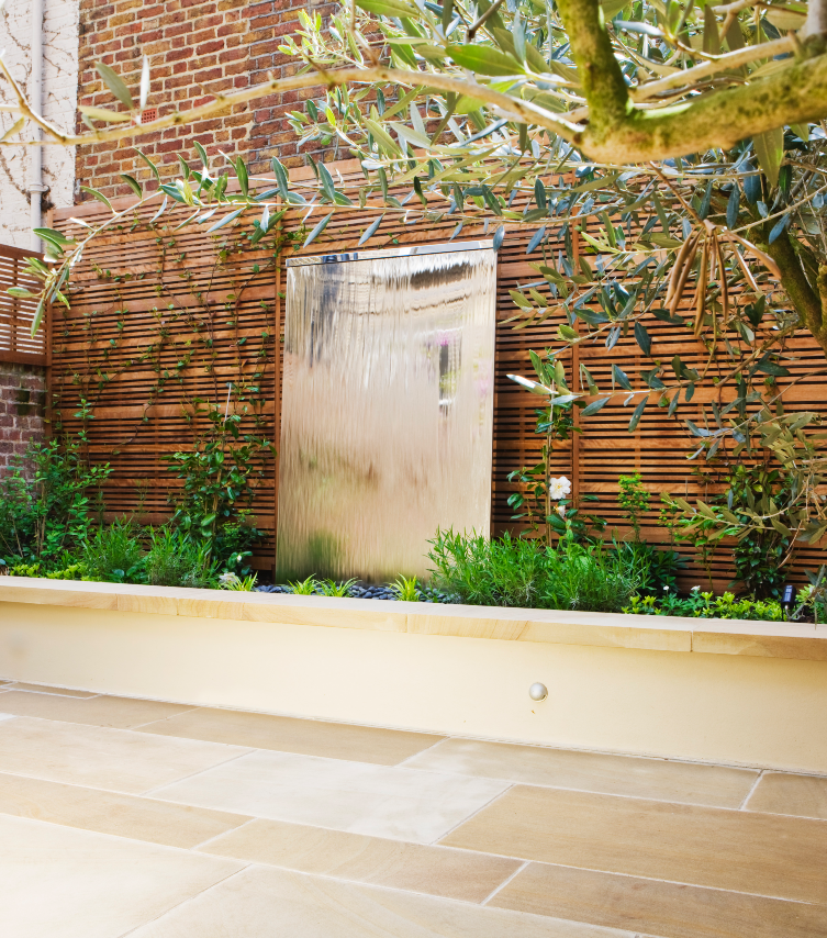 Mirror-polished stainless steel water wall in garden bed off of stone patio in modern backyard with wooden fence