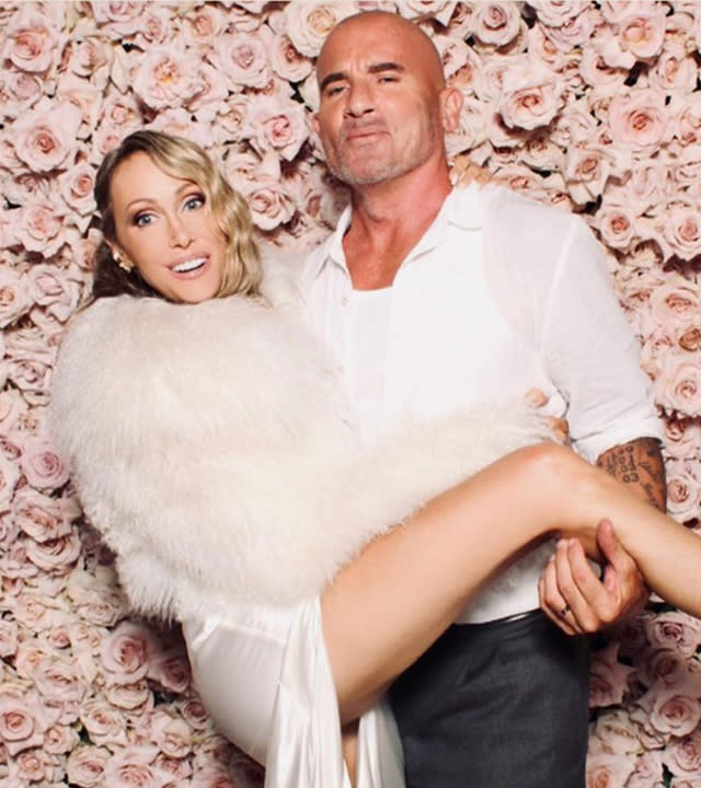 Tish Cyrus being held by Dominic Purcell in front of. a wall of pink flowers