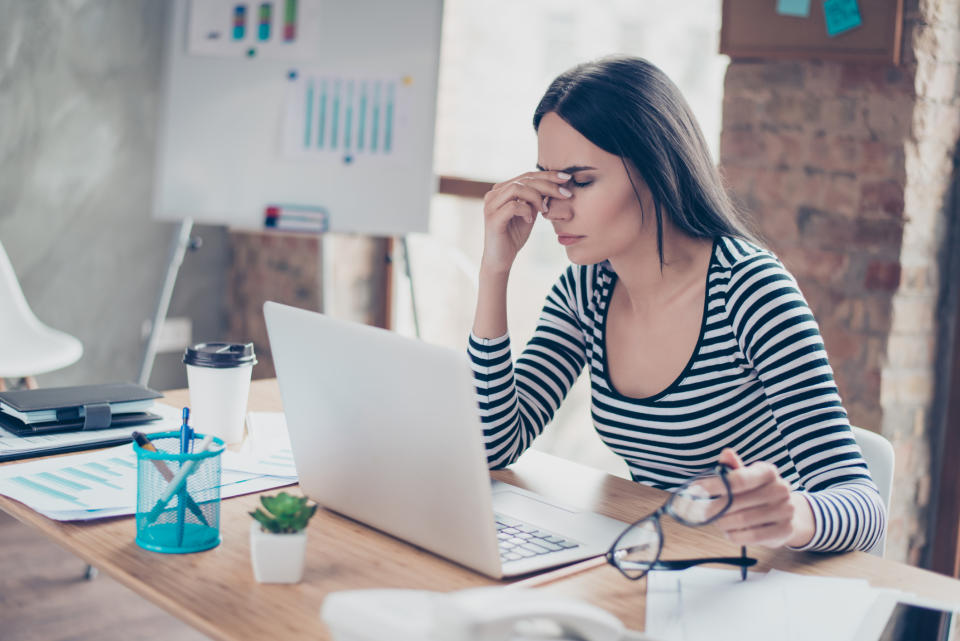 Woman at laptop holding a pair of glasses in one hand and pinching between her eyes with the other