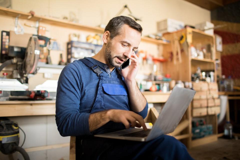 A man looks at his laptop's screen while on the phone in a workshop.