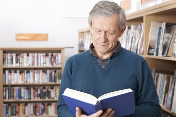 Older man reading a book while standing next to shelves of books