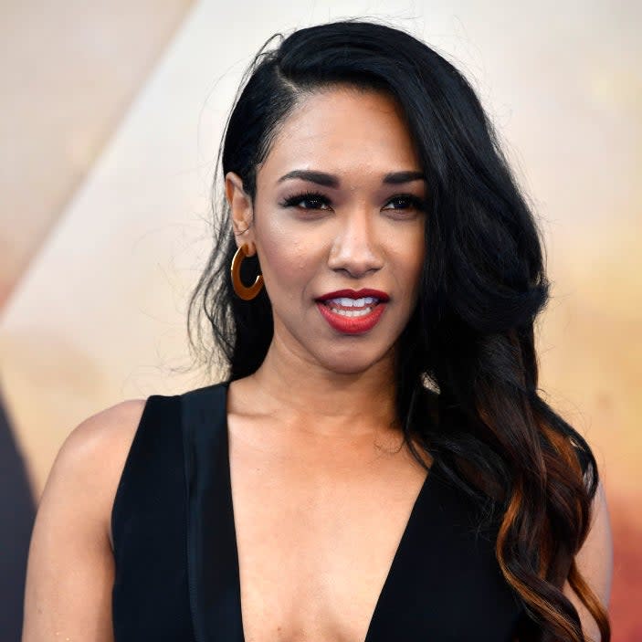 Candice Patton on the red carpet wearing a sleek, sleeveless black dress with gold hoop earrings and wavy hair styled to one side