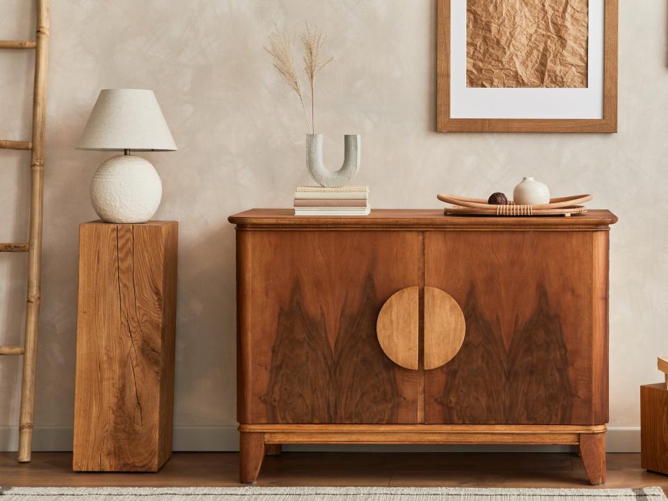 A wooden cabinet with brown picture frames and abstract art, a simple end table with a white lamp, and a decorative ladder against a wall