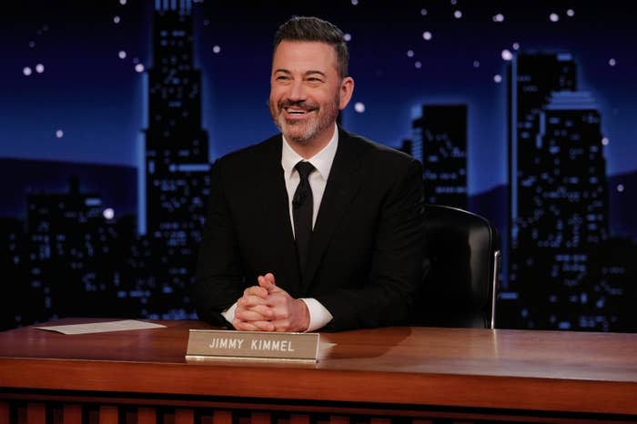 Jimmy Kimmel sits at a desk with a nameplate reading "Jimmy Kimmel," smiling in a dark suit and tie on a nighttime talk show set