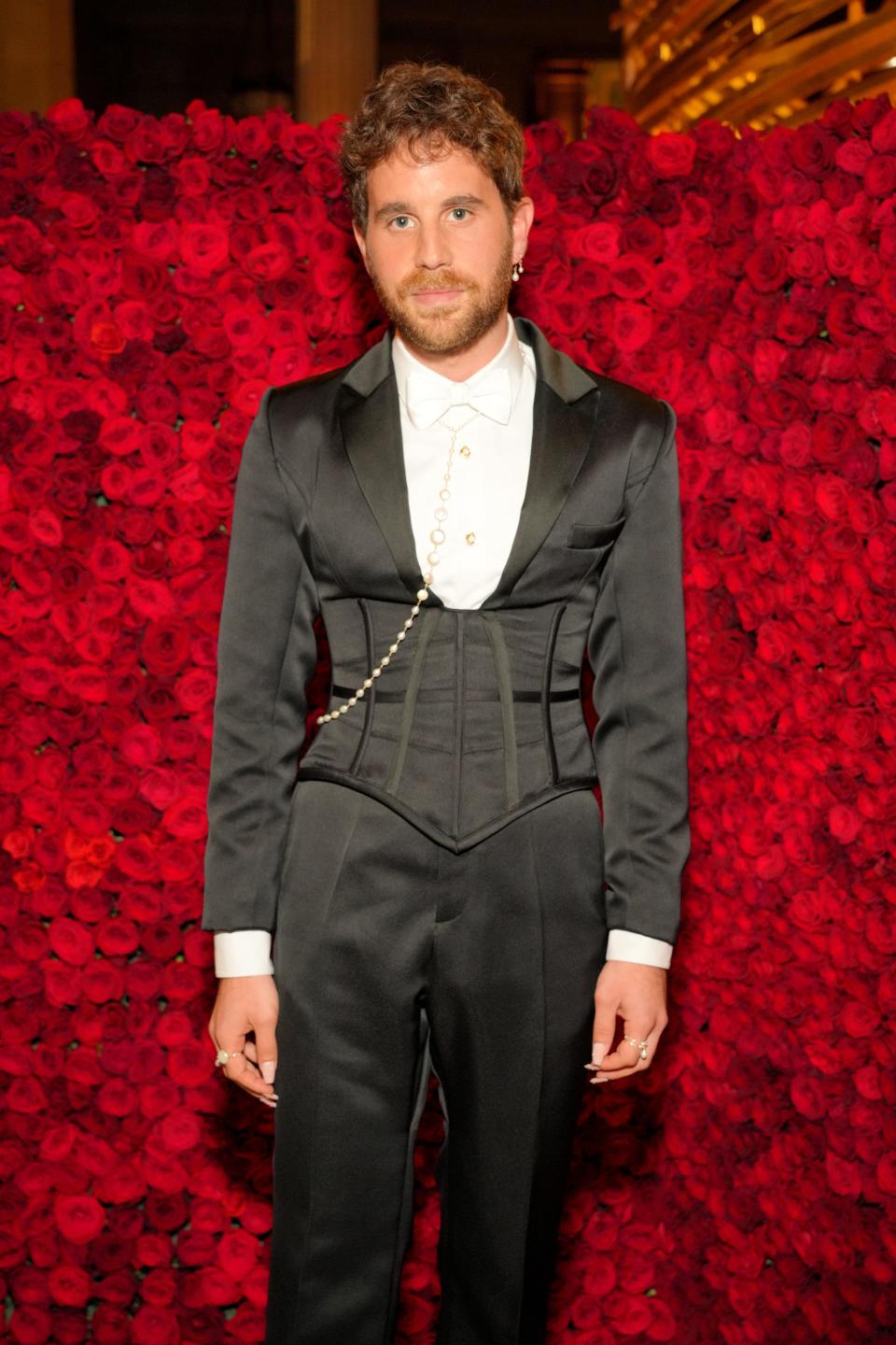 Ben in a black tuxedo with corset detailing around the waist of his jacket. There's a strand of pearls attached to his white button-down and bow tie. He's wearing black trousers. He has a long white manicure.