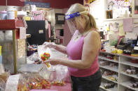 In this Thursday, May 28, 2020, photo, Susan Truax, owner of Bruce's Candy Kitchen, wears a protective plastic face shield while putting candy into a bag in Cannon Beach, Ore. With summer looming, Cannon Beach and thousands of other small, tourist-dependent towns nationwide are struggling to balance fears of contagion with their economic survival in what could be a make-or-break summer. (AP Photo/Gillian Flaccus)