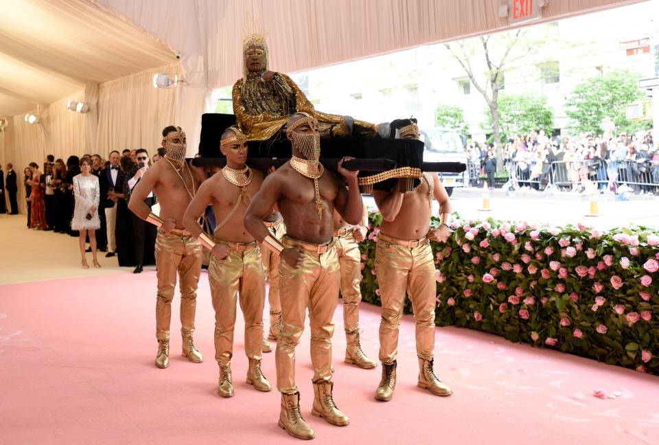 Billy Porter arrives at the Met Gala in an elaborate gold look, carried in by six men in gold pants and headpieces.