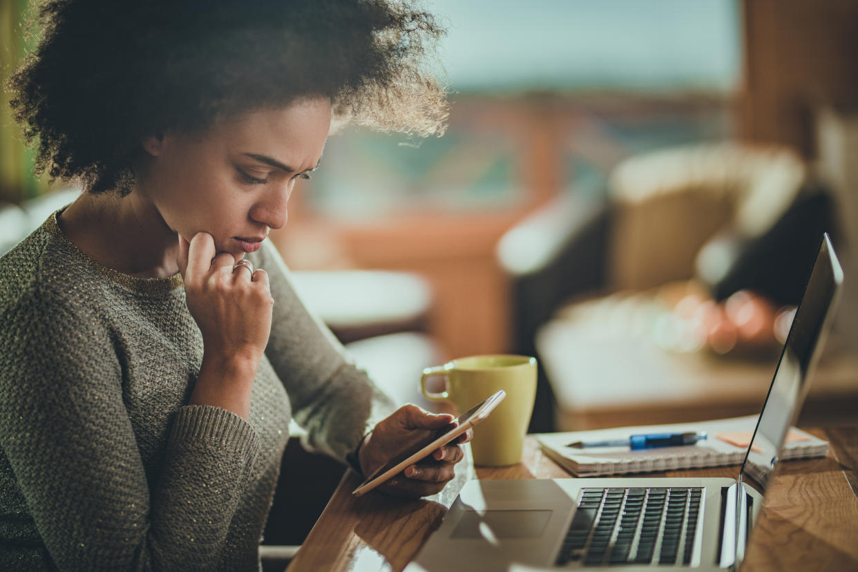 A young Black woman frowns as she looks down at her phone while sitting at a table in front of her laptop on age timeline