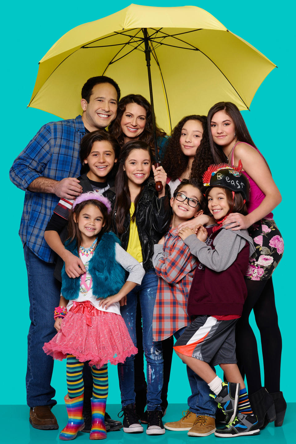 Nine people under a yellow umbrella, dressed in casual and playful clothing, smiling and posing closely together; image on a turquoise background