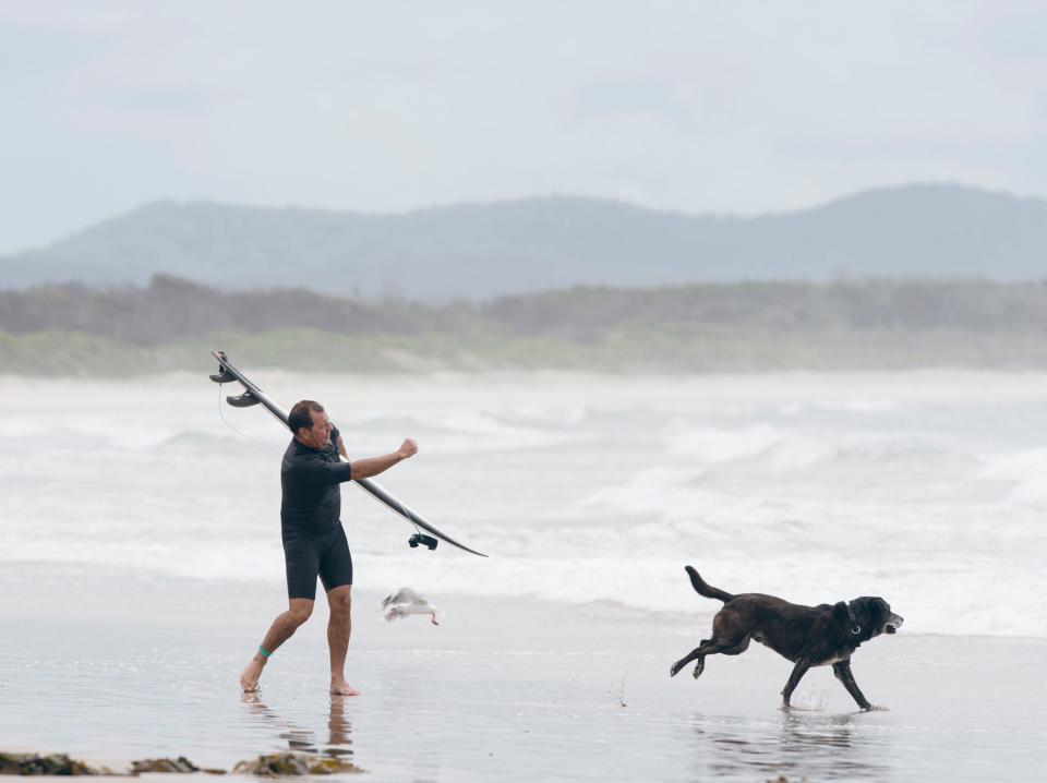 A photo of a man on the beach with a surfboard over his head, following a running dog.