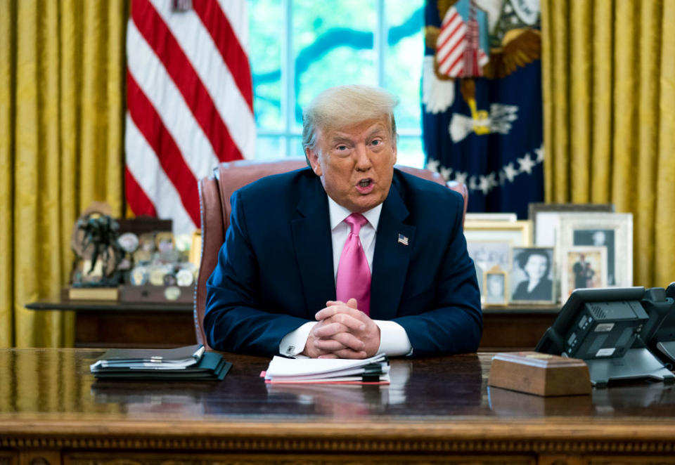 Donald Trump seated at the Oval Office desk, hands clasped, with flags and framed photos in the background