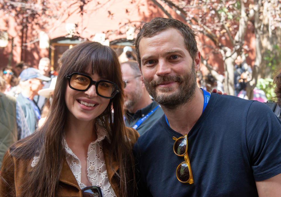 Dakota Johnson and Jamie Dornan smiling at an outdoor event. Dakota is wearing oversized glasses and a lace top. Jamie is in a casual T-shirt