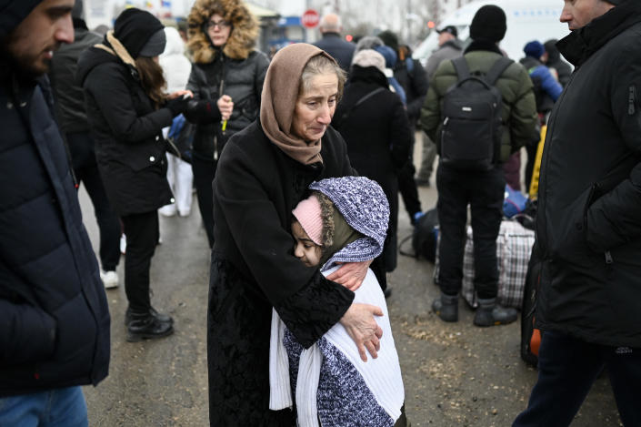 An older woman in a scarf looking tearful hugs a small girl, as refugees in winter clothes, some in backpacks, stand waiting.