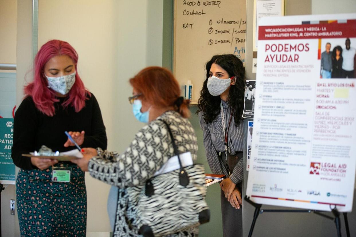 Two women, both masked, speak while looking over a clipboard as another woman, also masked, stands nearby