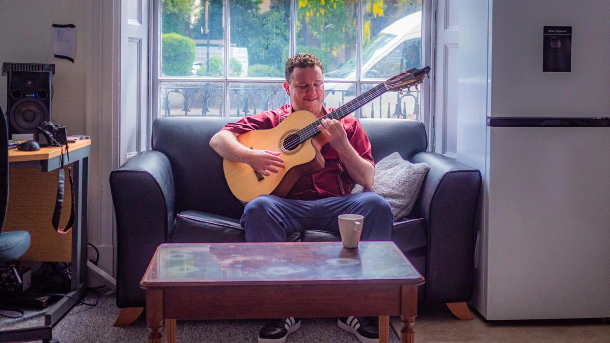 Eddy playing the guitar while sitting on a sofa next to a window, with a coffee table in front of him