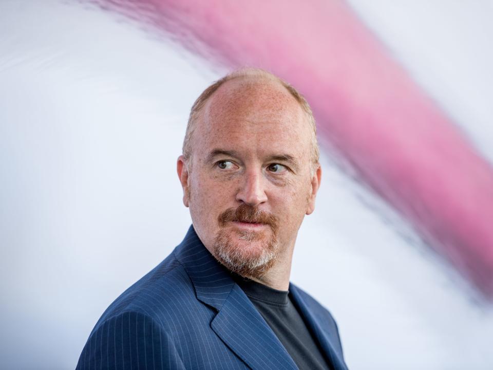 Louis C.K. stands on a red carpet.