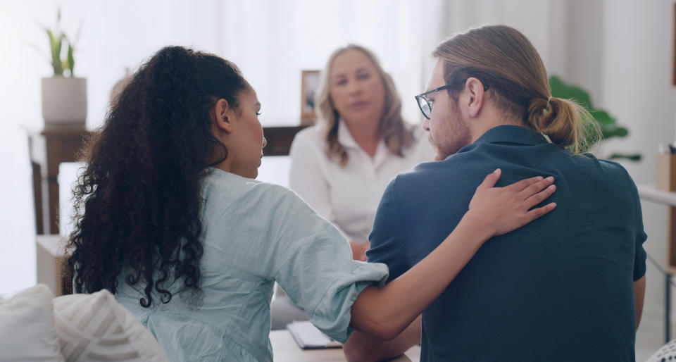 A woman with long, black curly hair places her hand on a man's back to comfort him as he turns to speak to her, with another woman in the background listening to them