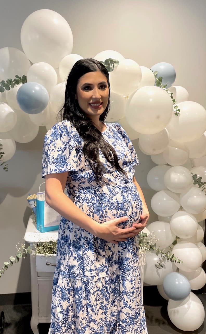 pregnant woman with long brown hair standing in front of white balloons holding baby bump wearing white and blue floral maxi dress