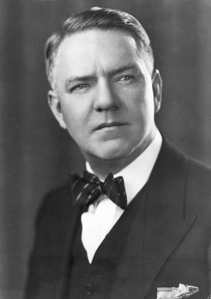 A man in formal attire with a suit and bow tie stares off to the side with a neutral expression