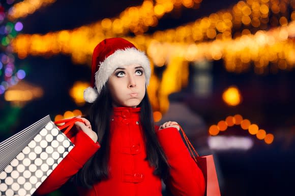 A woman puffs out her cheeks and looks upward while wearing a Santa hat and holding shopping bags.