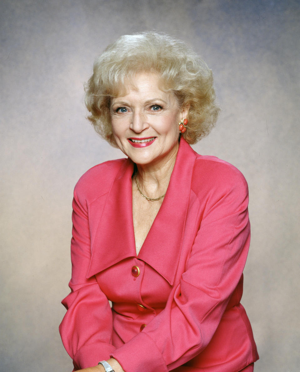 Betty White pose en souriant dans un blazer rose boutonné avec revers et boucles d'oreilles assorties