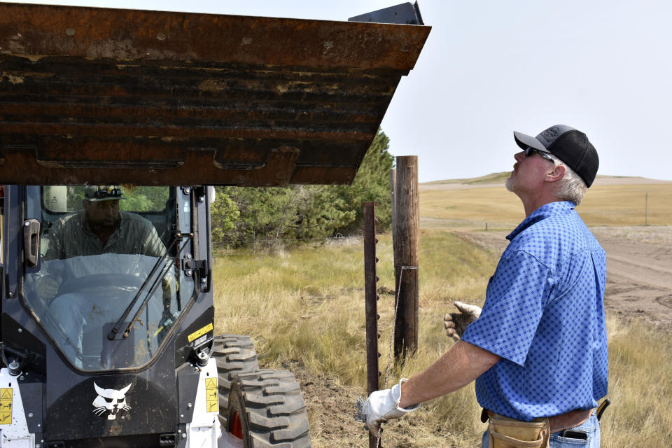 Howdy Lawlar, right holds a steel post as Charlie Lewis, left prepares to use a Bobcat to push it into the ground as they repair a barbed wire fence on Lawlar's farm, Aug. 25, 2021, in McKenzie County, N.D. Lewis came to the county to work in the booming oil fields but now works as Lawlar's farmhand. (AP Photo/Matthew Brown)