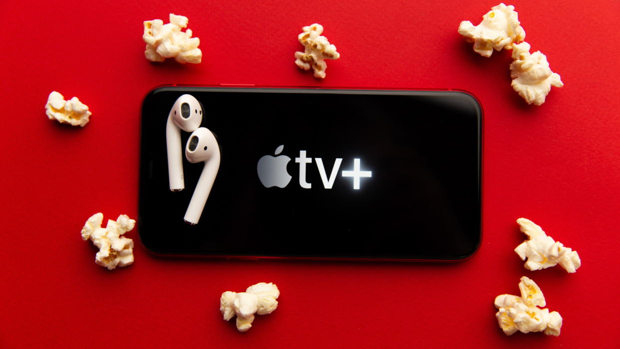  IPhone with Apple TV Plus logo, a pair of AirPods on top, laying on a red background with pieces of popcorn. 