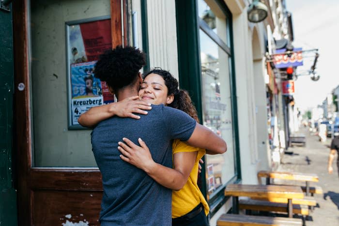 A couple hugs outside a building on a street, with a poster in the background advertising an event in Kreuzberg. The scene suggests a warm, affectionate moment