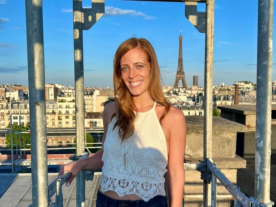 Amy Kehrig, a white woman in a white tank top, poses on a Paris rooftop with the Eiffel Tower in the background.