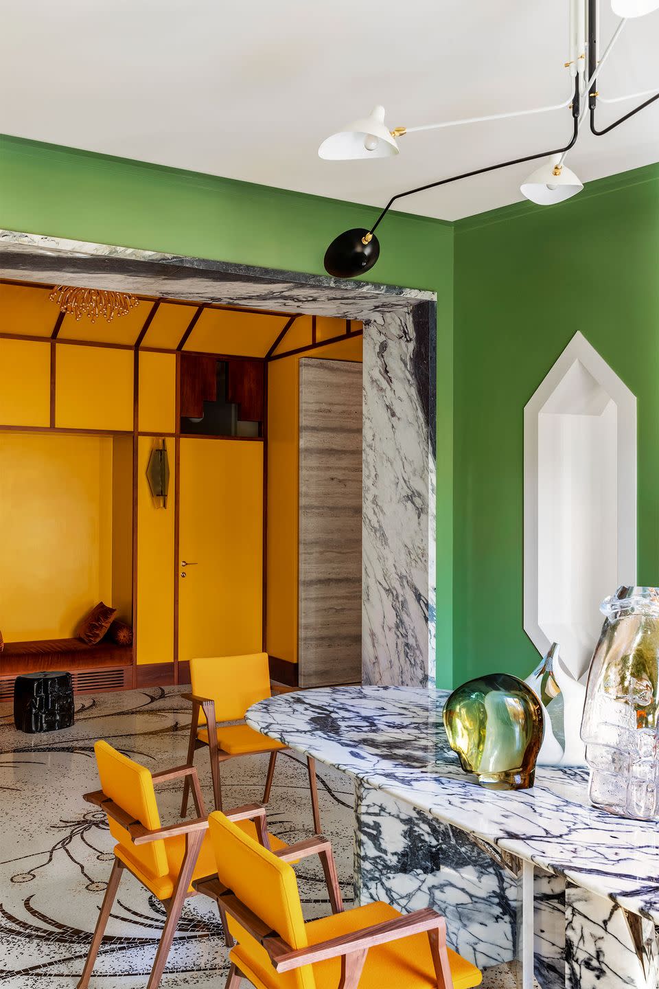 dining room with green walls, white and black marble table and chairs, white and black swirl patterned floor, goldenrod colored chairs, glass sculptures on table, five armed chandelier, interior hexagonal windows