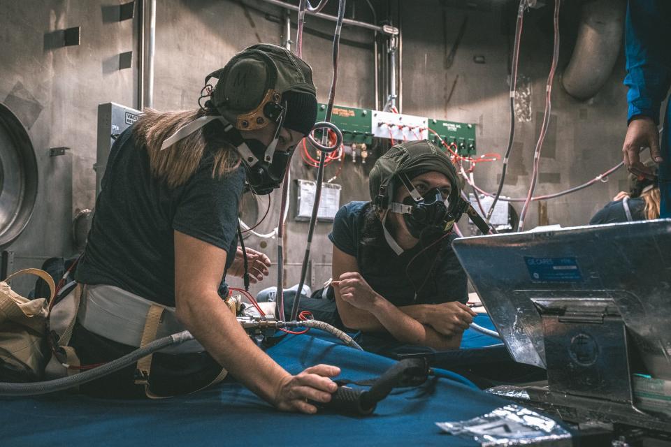 two people wearing large, bulky oxygen face masks sitting in a concrete room on blue mats