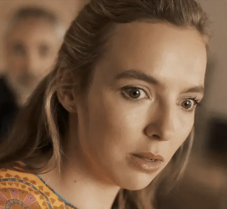 Jodie Comer looks surprised and focused, her hair pulled back, wearing a patterned top, with a blurred person in the background