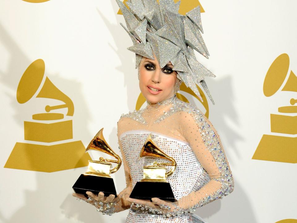 Gaga with dark eyeliner around her eyes, a silver spiked headpiece, and a silver dress holding two Grammy awards.