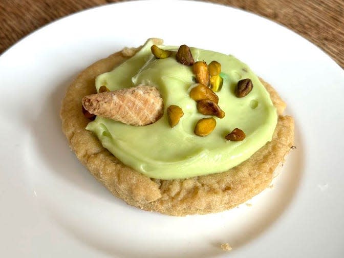 A Crumbl cookie with green frosting, a small cone, and pistachios on a white plate. The plate is on a wooden table.