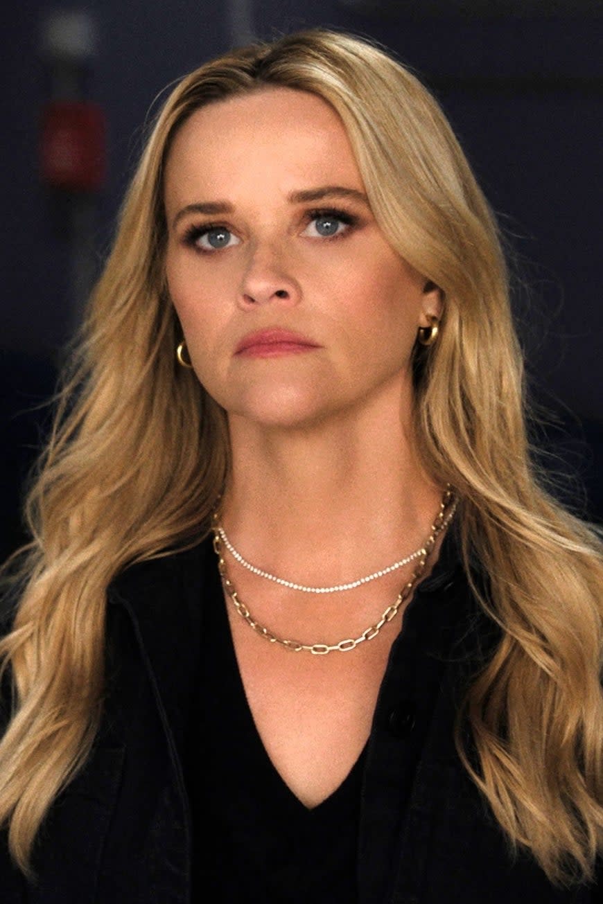 Reese Witherspoon stands in a hallway, looking serious, wearing a black outfit with layered necklaces