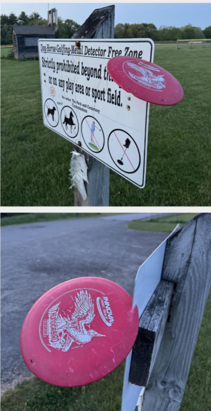 Two red frisbees stuck into a farm sign that prohibits dogs, horses, golfing, and metal detectors beyond certain points. Outdoor field in the background