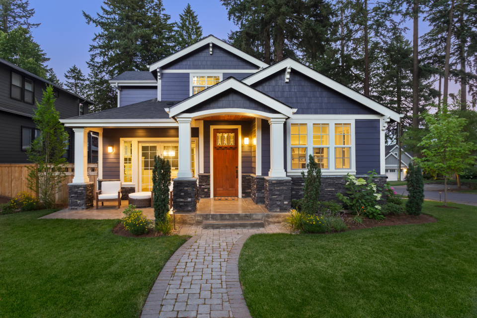 A blue house next to a wooded lot with its front porch lights turned on.