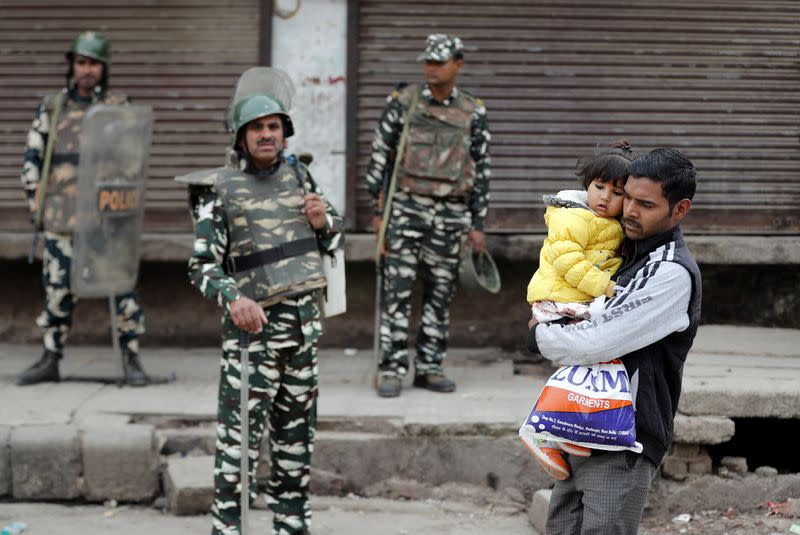 A man carrying a child walks past security forces in a riot affected area following clashes between people demonstrating for and against a new citizenship law in New Delhi