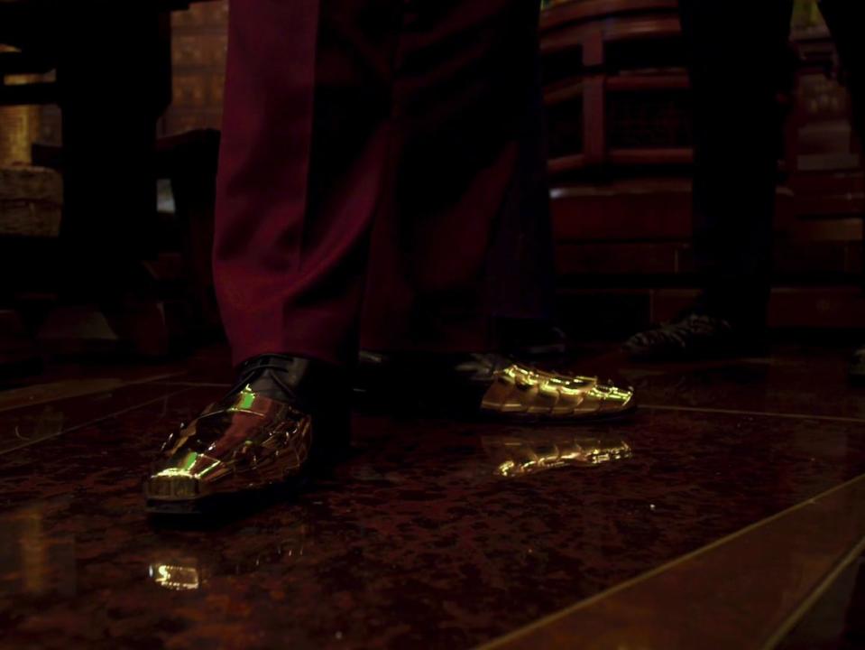 the feet of an adult man, glad in dress shoes that are plated with gold on the front. the man's leg is wearing a maroon pant, and he's standing on a shining floor.