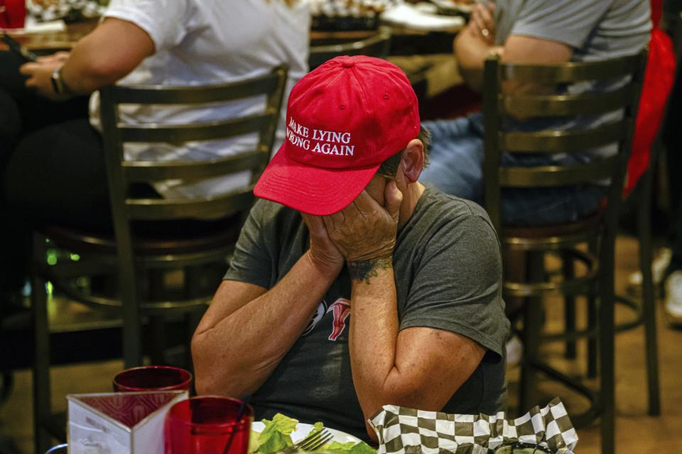 Person with face covered by hands wears a red hat with the text "MAKE LYING WRONG AGAIN" at a restaurant