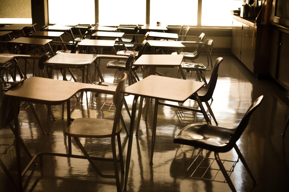 An empty classroom with rows of desks and chairs arranged in a typical school setting. Sunlight streams through the windows, reflecting on the shiny floor