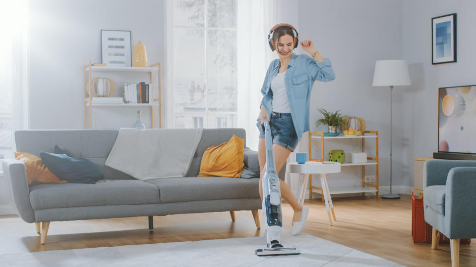 Shot of a Young Beautiful Woman in Jeans Shirt and Shorts Dancing and Vacuum Cleaning a Carpet in a Cozy Room at Home. She Uses a Modern Cordless Vacuum. She's Happy.