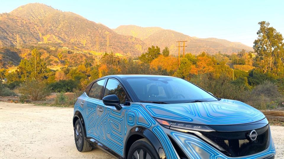 nissan ariya art car parked on a dirt road with trees and mountains in the background