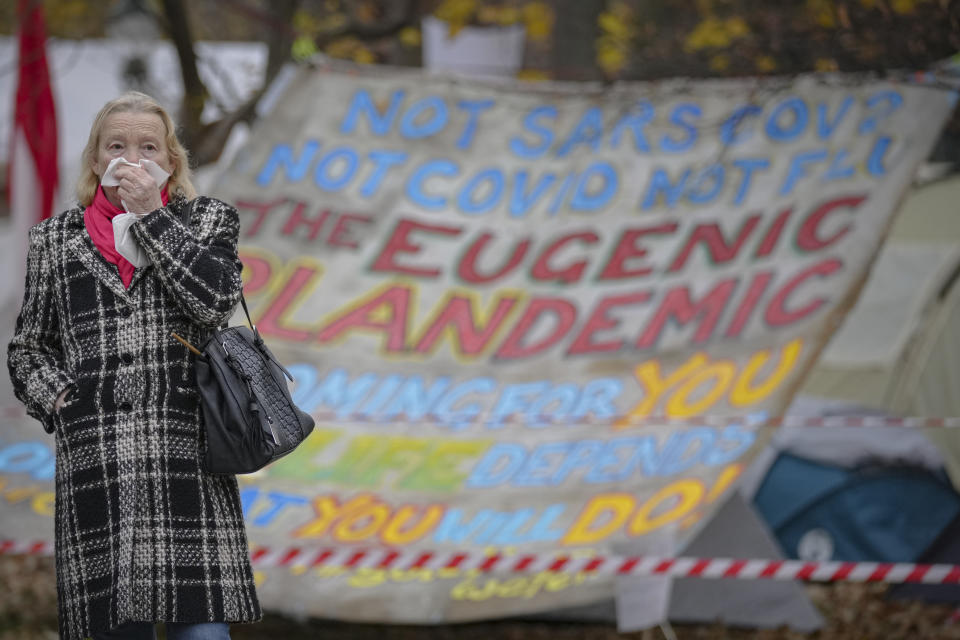 A woman covers her mouth and nose with a napkin as she walks by a small camp set up by opposers of the COVID-19 protection regulations in Vienna, Austria, Sunday, Nov. 21, 2021. The Austrian government announced a nationwide lockdown that will start Monday and comes as average daily deaths have tripled in recent weeks and hospitals in heavily hit states have warned that intensive care units are reaching capacity.(AP Photo/Vadim Ghirda)