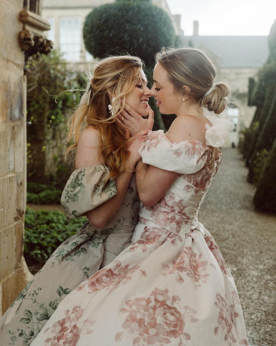 Two brides lean towards each other to kiss in front of a courtyard.