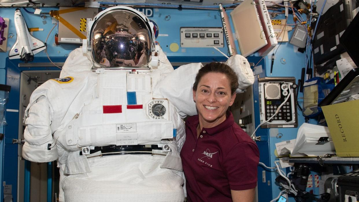  astronaut holds a spacesuit and poses next to it in the international space station. books and hardware are attached to the walls behind, including a model space shuttle 